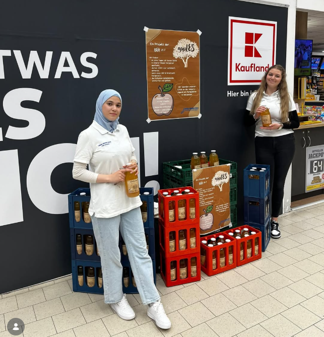 Zwei Sch&uuml;lerinnen stehen vor Apfelsaftkisten in einem Kaufland Supermarkt. Beide halten eine Flasche in der Hand. 