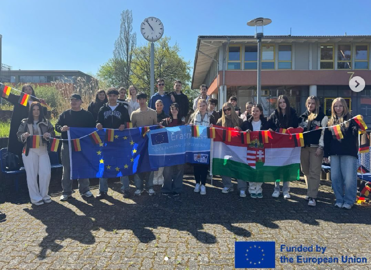 Gruppenbild: Jugendliche halten die deutsche, die ungarische und auf die Erasmus Flagge, auf einem Schulhof in der Sonne. Unten rechts steht auf dem Bild: "Funded by the European Union".