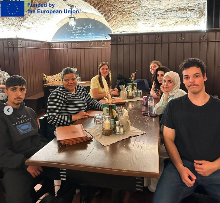 Eine Gruppe Schüler sitzt in einem Restaurant um einen Holztisch herum. Oben links steht das Logo "Funded by the European Union".