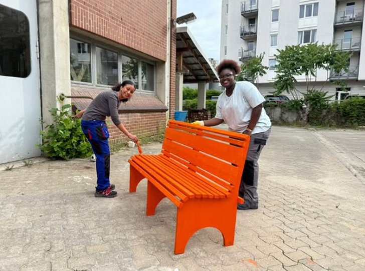 Zwei Schülerinnen streichen eine Holzbank im Freien in der Farbe Orange an. 