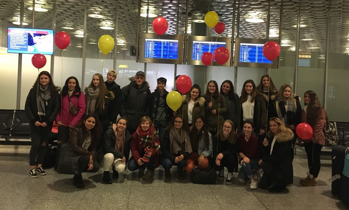 Gruppenbild: Jugendliche mit roten und gelben Luftballons in einer Flughafen-Wartehalle. 