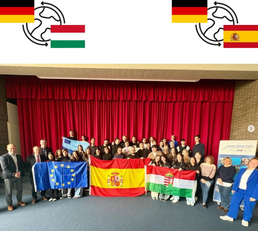 Eine Gruppe von Schülern steht vor einem roten Vorhang. Sie halten drei Fahnen hoch: die europäische, die spanische und die ungarische.  