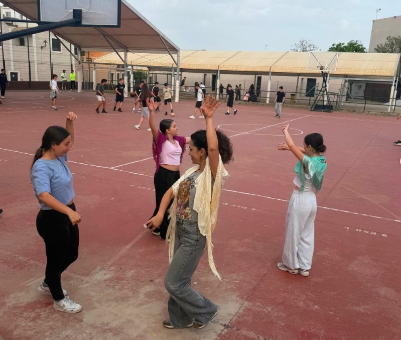 Schulfofszene: Im Vordergrund tanzen 4 spanische Mädchen Sevillanas (Flamenco), im Hintergrund spielen Kinder Basketball. 