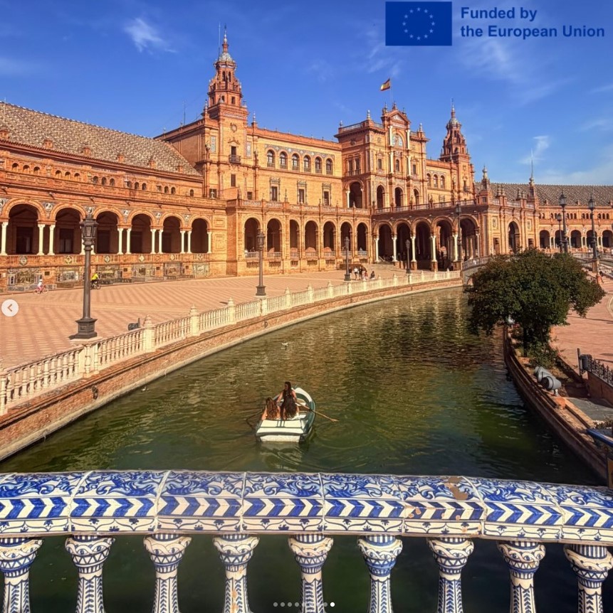Blick über ein ornamental verziertes Brückengeländer auf einen Fluss mit einem Boot und ein prachtvolles Sandsteingebäude mit vielen Verzierungen. Oben rechts steht: ‘Funded by the European Union’ mit EU-Flagge 
