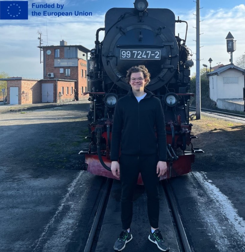 Ein jugendlicher Junge steht vor einer Lok. Oben links steht: &lsquo;Funded by the European Union&rsquo; mit EU-Flagge.