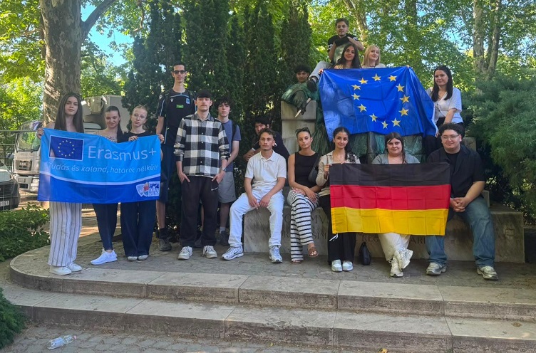 Ein schattiges Gruppenbild des Schüleraustauschs nach Ungarn. Die Schüler halten eine Erasmus-, eine Deutschland-, und eine Europa-Flagge hoch. 