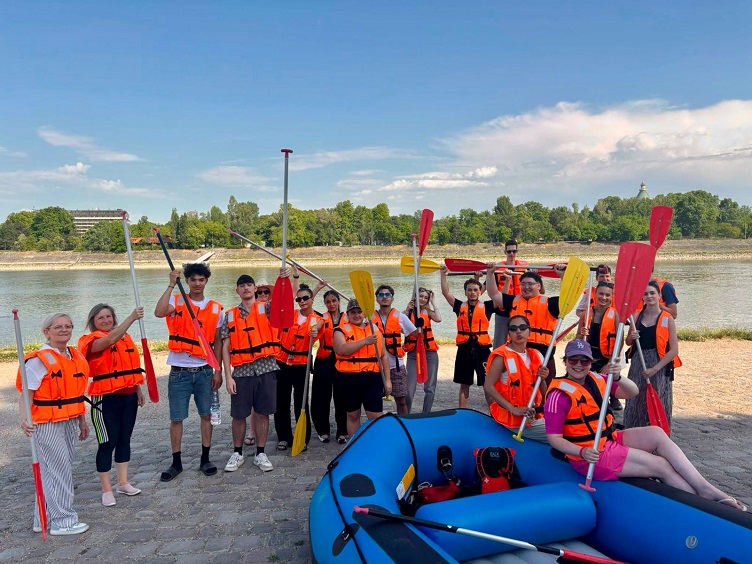 Ein Gruppenbild des Schüleraustauschs nach Ungarn: Viele Schüler in Schwimmwesten, und mit Paddeln, mit einem Schlauchboot am Strand. 