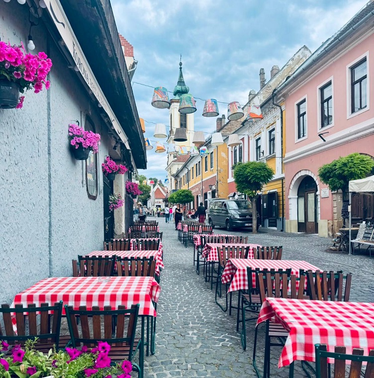 Eine historische, prunkvolle, Strasse mit Blumenschmuck und karierten Tischdecken in einem Cafe. Im Hintergrund ein Kirchturm.  