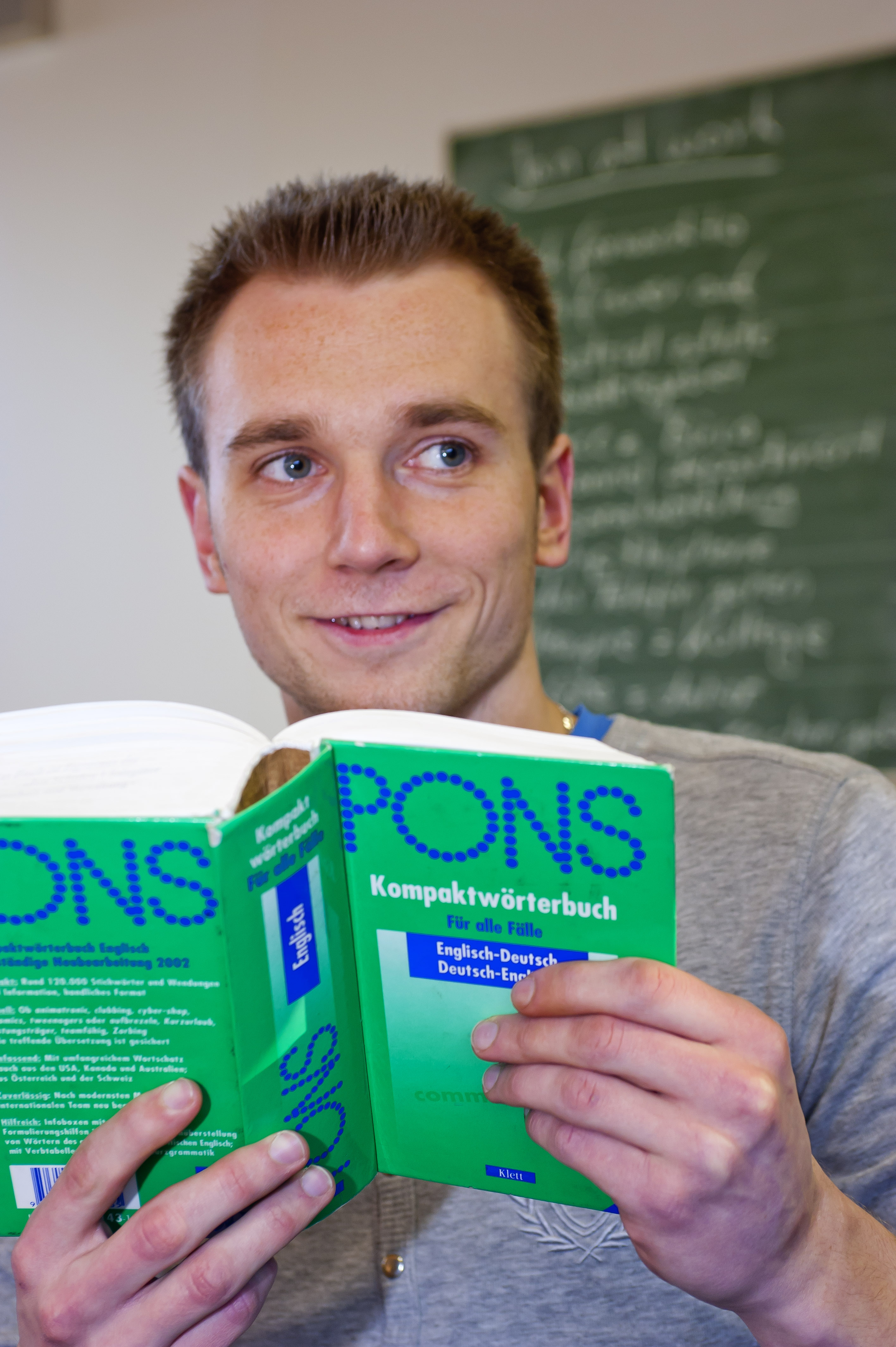 Ein Sch&uuml;ler schaut gut gelaunt &uuml;ber den Rand eines Fremdsprachenlexikons. 