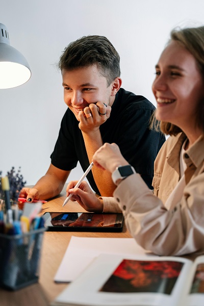Ein Sch&uuml;lerin und ein Sch&uuml;ler sitzen lachend am Tisch. Das M&auml;dchen hat ein Tablet vor sich liegen und einen Tablet-Stift in der Hand. 