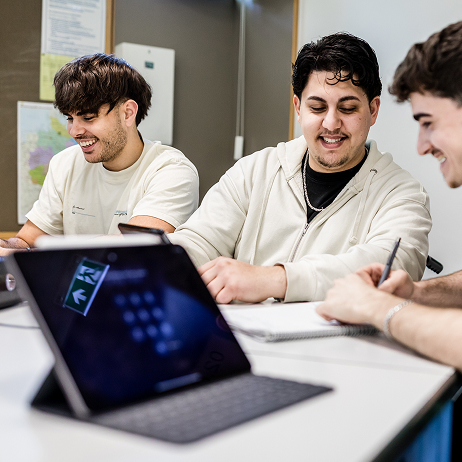 Drei jugendliche Sch&uuml;ler am Schultisch. Sie schreiben und lachen. Im Vordergrund steht ein Tablet. 