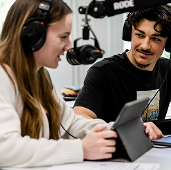 Zwei Sch&uuml;ler mit Kopfh&ouml;rern sprechen in ein Mikrofon. Das M&auml;dchen im Vordergrund liest von einem Tablet ab. 