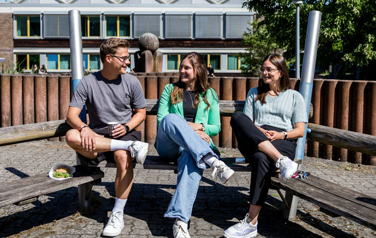 Drei Sch&uuml;ler sitzen lachend auf einer Bank in der Sonne. 