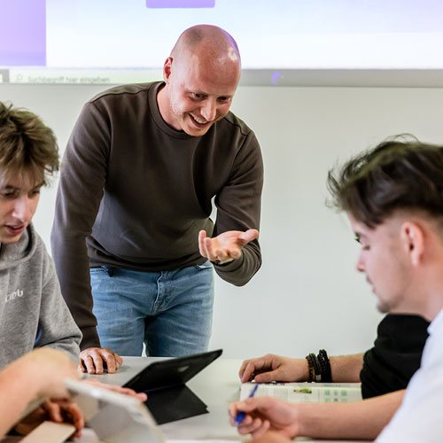 Ein Lehrer erkl&auml;rt lachend ein paar Sch&uuml;lern die am Tisch sitzen und schreiben etwas. Auf dem Tisch stehen Tablets.  