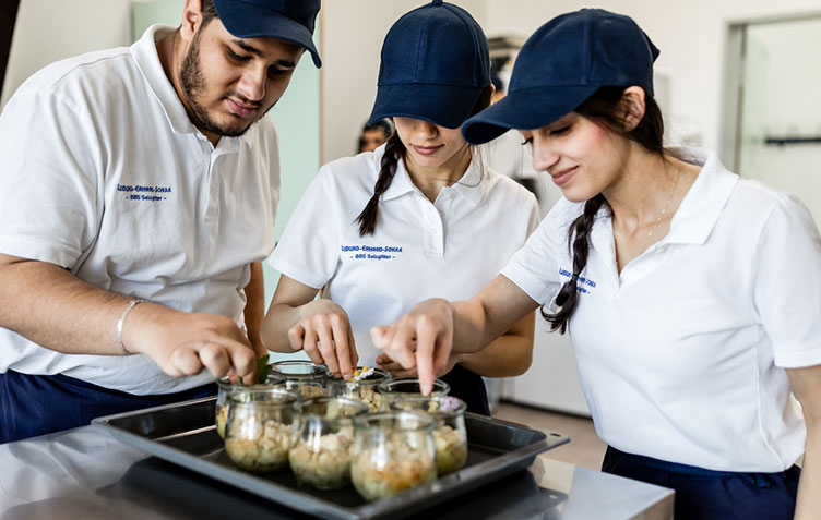 Drei Sch&uuml;ler in K&uuml;chenuniform steht vor zubereiteten Speisen in kleinen Glasf&ouml;rmchen. Sie schauen fr&ouml;hlich in die Gl&auml;ser. 