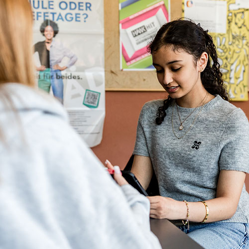 Blick &uuml;ber die Schulter einer Sch&uuml;lerin: Ihr gegen&uuml;ber ein M&auml;dchen am Tisch sitzend, und nach unten blickend.  